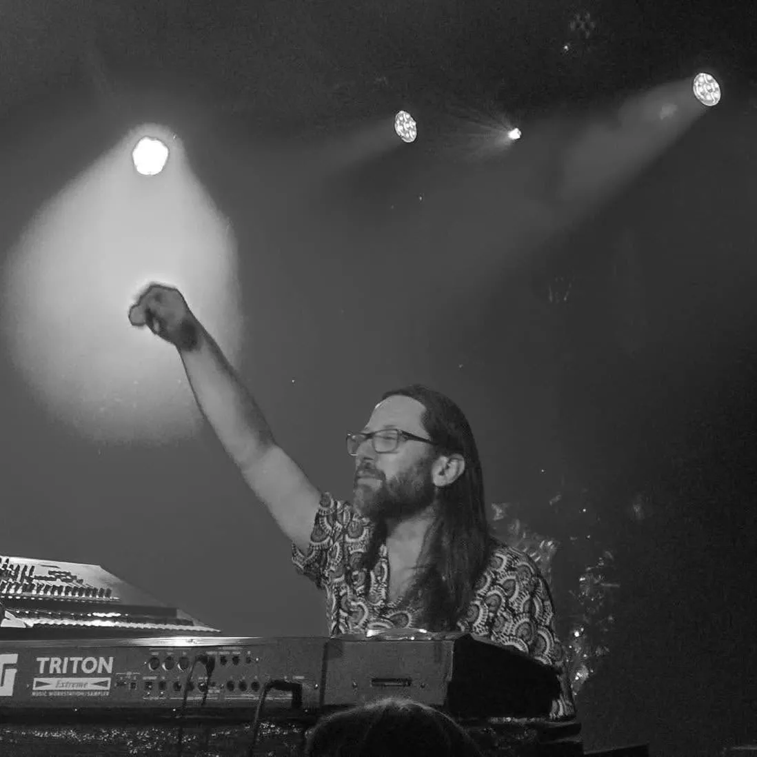 Black and white photo: a long-haired keyboard player in glasses raises one arm under stage lights, behind a Korg Triton Extreme