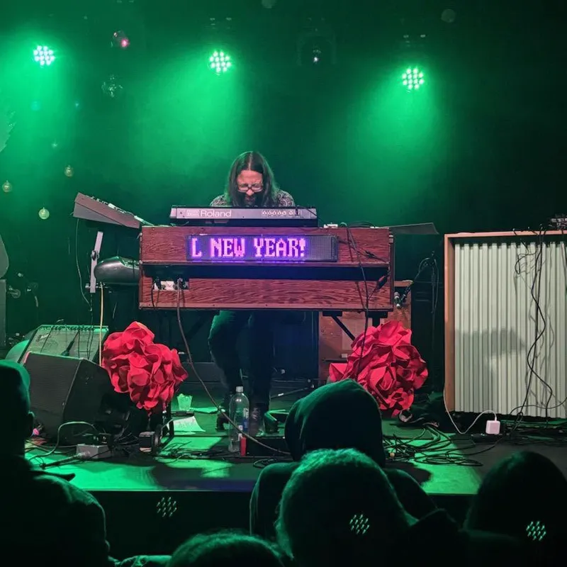 A keyboard player behind a wooden organ under green stage light, a scrolling LED sign on the organ reading NEW YEAR, red paper flowers either side