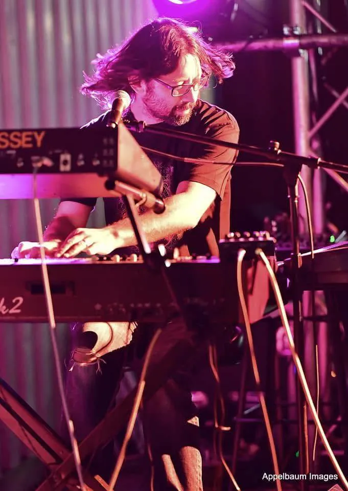 A long-haired keyboard player in glasses plays under magenta stage light, an ARP Odyssey above the keys