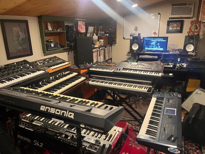 A low basement room packed with keyboards: an Ensoniq, a Hammond SK2, a Rhodes Mark II stage piano, an ARP Odyssey, Moog synths, monitors and a record shelf