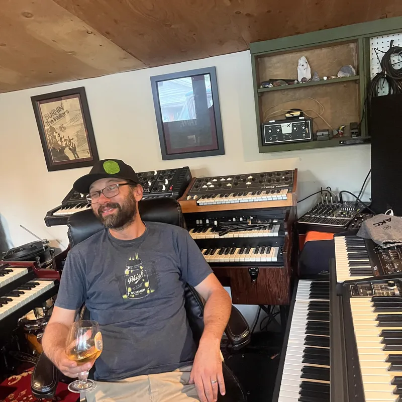 A smiling bearded man in glasses and a black cap sits in a leather chair holding a glass, surrounded by synthesizers and organs